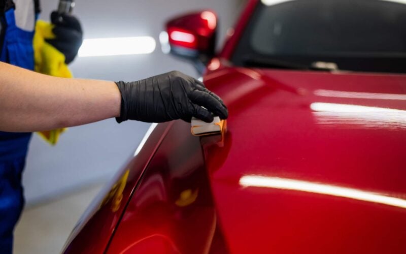 Gros plan d'un professionnel appliquant un traitement de protection céramique sur la carrosserie d'une voiture rouge.